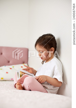 Young girl sitting on a bed reading a book, focusing on the story, promoting early learning and quiet childhood moments 140087464
