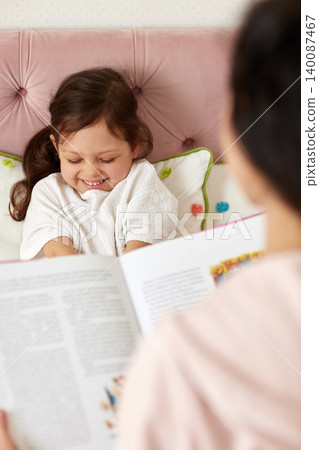Mother reading book to cheerful little daughter in bed, creating a warm moment of family bonding and care Mother reading book to cheerful little daughter in bed, creating a warm moment of family bonding and care 140087467