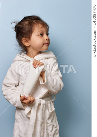 Child girl wearing a white bathrobe holding a blank cream tube on a blue background 140087476