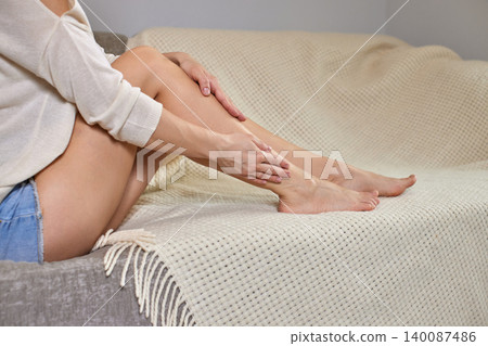 Woman sitting on a sofa massaging her shin and calf to relieve muscle pain, cramp, or exhaustion after exercise Woman sitting on a sofa massaging her shin and calf to relieve muscle pain, cramp, or exhaustion after exercise 140087486