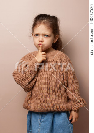 Young child girl with finger on lips asking for quiet with thoughtful expression on beige background Young child girl with finger on lips asking for quiet with thoughtful expression on beige background 140087526