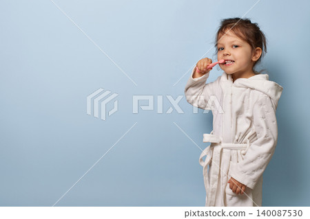 Child girl in white bathrobe practicing brushing teeth against a clean blue background, copy space 140087530