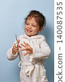 happy child girl in a white bathrobe learning self-care with nail scissors 140087535