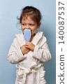 Little child girl in a white bathrobe holding a hairbrush to her nose, looking away on blue background 140087537
