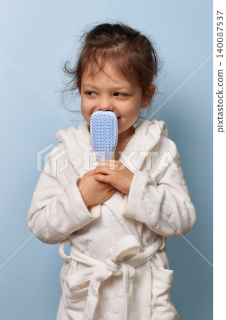 Little child girl in a white bathrobe holding a hairbrush to her nose, looking away on blue background Little child girl in a white bathrobe holding a hairbrush to her nose, looking away on blue background 140087537