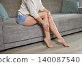 Woman on sofa applying moisturizer to legs, self-massage for soft, hydrated skin and relaxed at-home beauty and wellness routine 140087540