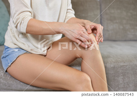 young woman experiencing knee pain, gently touching and massaging her painful leg joint, finding relief from discomfort or injury 140087543