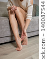 Woman sitting on sofa massaging cream into foot, moisturizing skin for soft, healthy feet during a relaxed home self-care routine 140087544