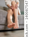 Woman sitting on a sofa massaging her feet and calves, experiencing comfort and relief from leg pain and muscle fatigue 140087547