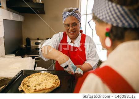 Smiling Mature Hispanic Woman Baking Pizza in a Bright Community Kitchen with a Female Friend Smiling Mature Hispanic Woman Baking Pizza in a Bright Community Kitchen with a Female Friend 140088112
