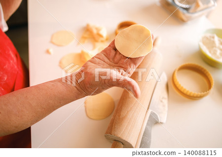 Elderly Caucasian woman hands making circular dough for cookies in a sunlit domestic kitchen Elderly Caucasian woman hands making circular dough for cookies in a sunlit domestic kitchen 140088115