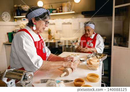 Active senior Caucasian woman rolls fresh dough with a rolling pin in a professional bakery kitchen. Active senior Caucasian woman rolls fresh dough with a rolling pin in a professional bakery kitchen. 140088125