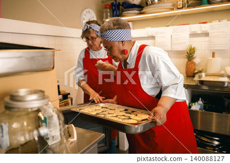 Active senior Black and White women carrying fresh pastries in a commercial community kitchen Active senior Black and White women carrying fresh pastries in a commercial community kitchen 140088127