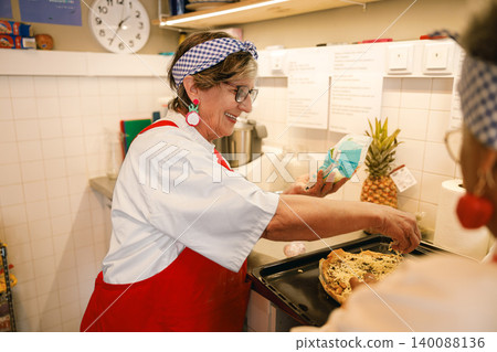 Happy senior Caucasian female chef preparing fresh food on a baking sheet in a sunny indoor kitchen Happy senior Caucasian female chef preparing fresh food on a baking sheet in a sunny indoor kitchen 140088136