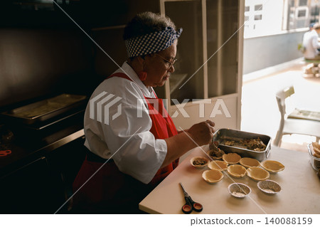 Senior Black woman chef filling pastry shells in a commercial kitchen with focused precision Senior Black woman chef filling pastry shells in a commercial kitchen with focused precision 140088159