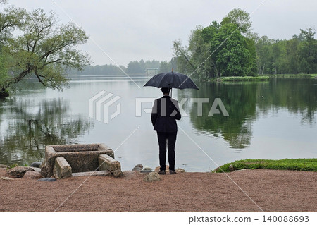 Rainy day, lone figure stands by calm lake, view from the back. Rainy day, lone figure stands by calm lake, view from the back. 140088693