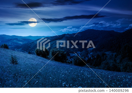 mountainous countryside landscape in summer at night. full moon over grassy field on a hillside. view of a forested mountain ridge in the distance. mysterious weather with cloudy sky 140089296