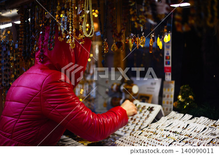 Woman in a bright red puffer jacket browsing an amber jewelry stall at a night market Woman in a bright red puffer jacket browsing an amber jewelry stall at a night market 140090011
