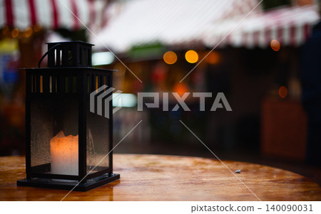 Black metal lantern with a lit candle on a wet wooden table at a festive market Black metal lantern with a lit candle on a wet wooden table at a festive market 140090031