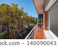 A sunny balcony view overlooking a tree-lined street in an urban area. Bright blue sky and modern buildings are visible 140090043