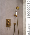 A modern shower fixture with a gold finish and a handheld showerhead. The wall has a smooth gray texture with white tiles in the background. 140090050