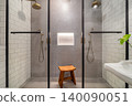 Modern bathroom with two glass shower stalls, a wooden stool, and a small green plant. The walls are tiled in white and gray tones. 140090051
