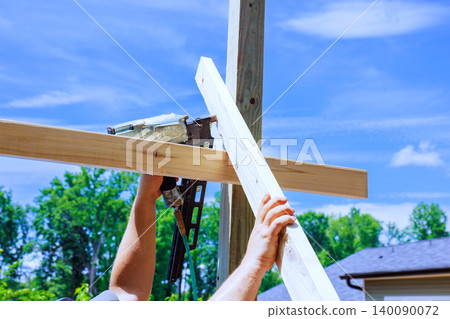 Worker is working on construction project by using nail gun to secure wooden beams against post in yard. Worker is working on construction project by using nail gun to secure wooden beams against post in yard. 140090072