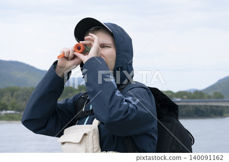 Man looking through a small telescope at a river landscape, demonstrating travel gear in real use during outdoor exploration. 140091162