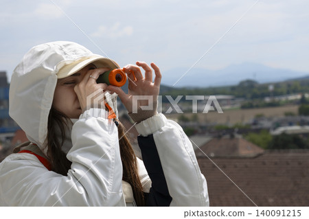 Young woman looking through a small orange telescope outdoors, showcasing a portable magnification device for everyday use. 140091215