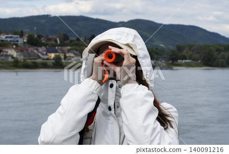 Woman in a hooded jacket looking through a small telescope at mountains, illustrating the use of a compact optical device. 140091216