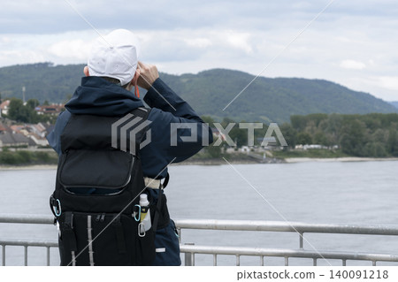 Man with backpack looking at a river and village through a small orange telescope, capturing a practical field observation moment. 140091218