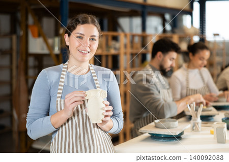 Girl in apron stands with vase in hands, finished clay products from craftsperson Girl in apron stands with vase in hands, finished clay products from craftsperson 140092088