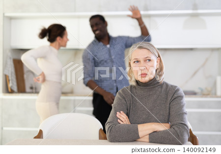 Upset mature woman sitting at table while married couple quarreling in background Upset mature woman sitting at table while married couple quarreling in background 140092149
