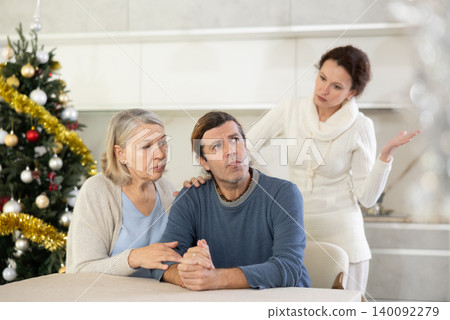 Woman is actively sorting out her relationship with man, and elderly woman is calming man while sitting at table in kitchen near christmas tree Woman is actively sorting out her relationship with man, and elderly woman is calming man while sitting at table in kitchen near christmas tree 140092279