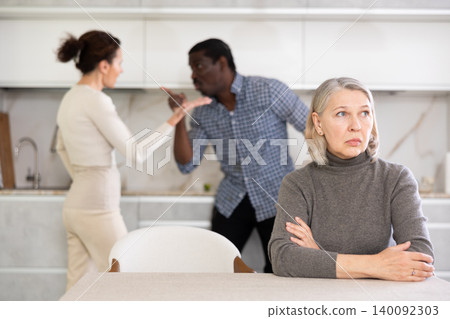 Elderly sad woman sits at table during family quarrel Elderly sad woman sits at table during family quarrel 140092303