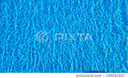 Real swimming pool water photograph featuring bright sunlight reflections, blue tiles, and dynamic ripple patterns. 140092887