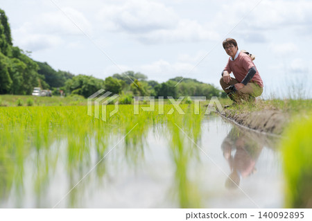 一位農民蹲在稻田邊,面帶微笑。 一位農民蹲在稻田邊,面帶微笑。 140092895