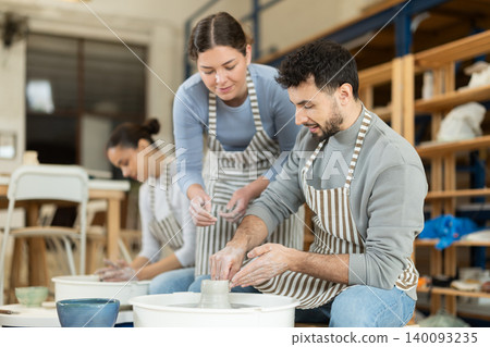 Man craftsperson busy molding clay products using pottery wheel in lesson with girl teacher Man craftsperson busy molding clay products using pottery wheel in lesson with girl teacher 140093235