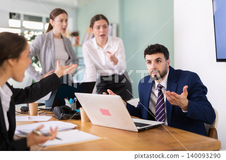 Frustrated man sits at a table with a laptop in front of his colleagues in office Frustrated man sits at a table with a laptop in front of his colleagues in office 140093290
