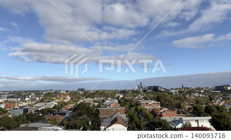 A wide-angle panoramic view of the suburban residential neighborhood with distinctive red roofs, looking towards the distant city CBD skyline under a bright blue sky with scattered clouds. A wide-angle panoramic view of the suburban residential neighborhood with distinctive red roofs, looking towards the distant city CBD skyline under a bright blue sky with scattered clouds. 140093716