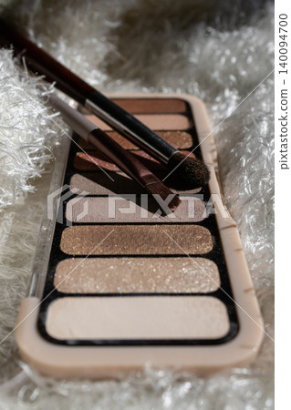 Eyeshadow palette displaying a range of neutral and glittering shades with makeup brushes on a soft, textured surface, representing beauty and cosmetic application Eyeshadow palette displaying a range of neutral and glittering shades with makeup brushes on a soft, textured surface, representing beauty and cosmetic application 140094700