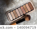 Eyeshadow palette displaying a range of neutral nude and glitter shades for everyday makeup looks, next to an applicator brush on a soft white furry background 140094719