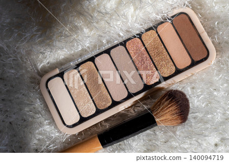 Eyeshadow palette displaying a range of neutral nude and glitter shades for everyday makeup looks, next to an applicator brush on a soft white furry background Eyeshadow palette displaying a range of neutral nude and glitter shades for everyday makeup looks, next to an applicator brush on a soft white furry background 140094719