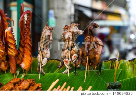 Exotic roasted street food skewers featuring giant octopus tentacles and frogs displayed on fresh banana leaf at outdoor market stall in Vietnam Exotic roasted street food skewers featuring giant octopus tentacles and frogs displayed on fresh banana leaf at outdoor market stall in Vietnam 140094823