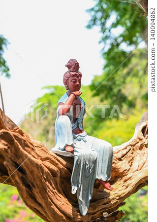 Beautiful Guanyin Bodhisattva statue sitting on driftwood branch surrounded by green trees in tropical Vietnam garden 140094862