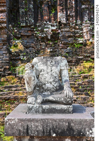 Ancient headless stone statue of Hindu deity on a pedestal in front of a red brick wall at My Son Sanctuary in Vietnam 140094874