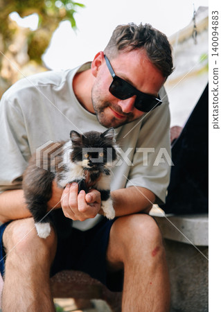 A caucasian 30 years old young man sitting outdoors and holding a cute fluffy domestic kitten in hands A caucasian 30 years old young man sitting outdoors and holding a cute fluffy domestic kitten in hands 140094883
