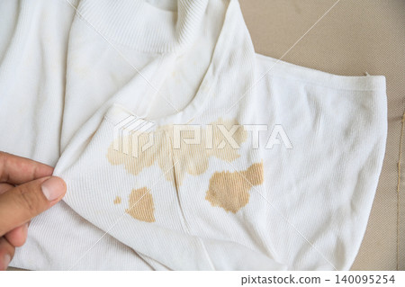 dirty yellow stains on a white rib-knit sleeveless shirt. Dirty clothes concept, sweat or food stains on fabric, laundry problem and stain removal or bleaching service background. dirty yellow stains on a white rib-knit sleeveless shirt. Dirty clothes concept, sweat or food stains on fabric, laundry problem and stain removal or bleaching service background. 140095254