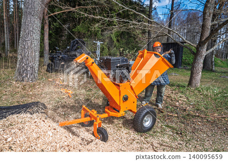 Woman operating wood chipper in forest, safety gear, wood chips flying, outdoor power equipment 140095659