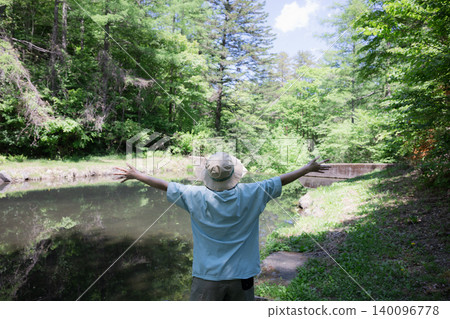 A woman takes a deep breath and spreads her arms while enjoying a forest bath; a faceless image showing her back as she enjoys the outdoors and summer camping. A woman takes a deep breath and spreads her arms while enjoying a forest bath; a faceless image showing her back as she enjoys the outdoors and summer camping. 140096778